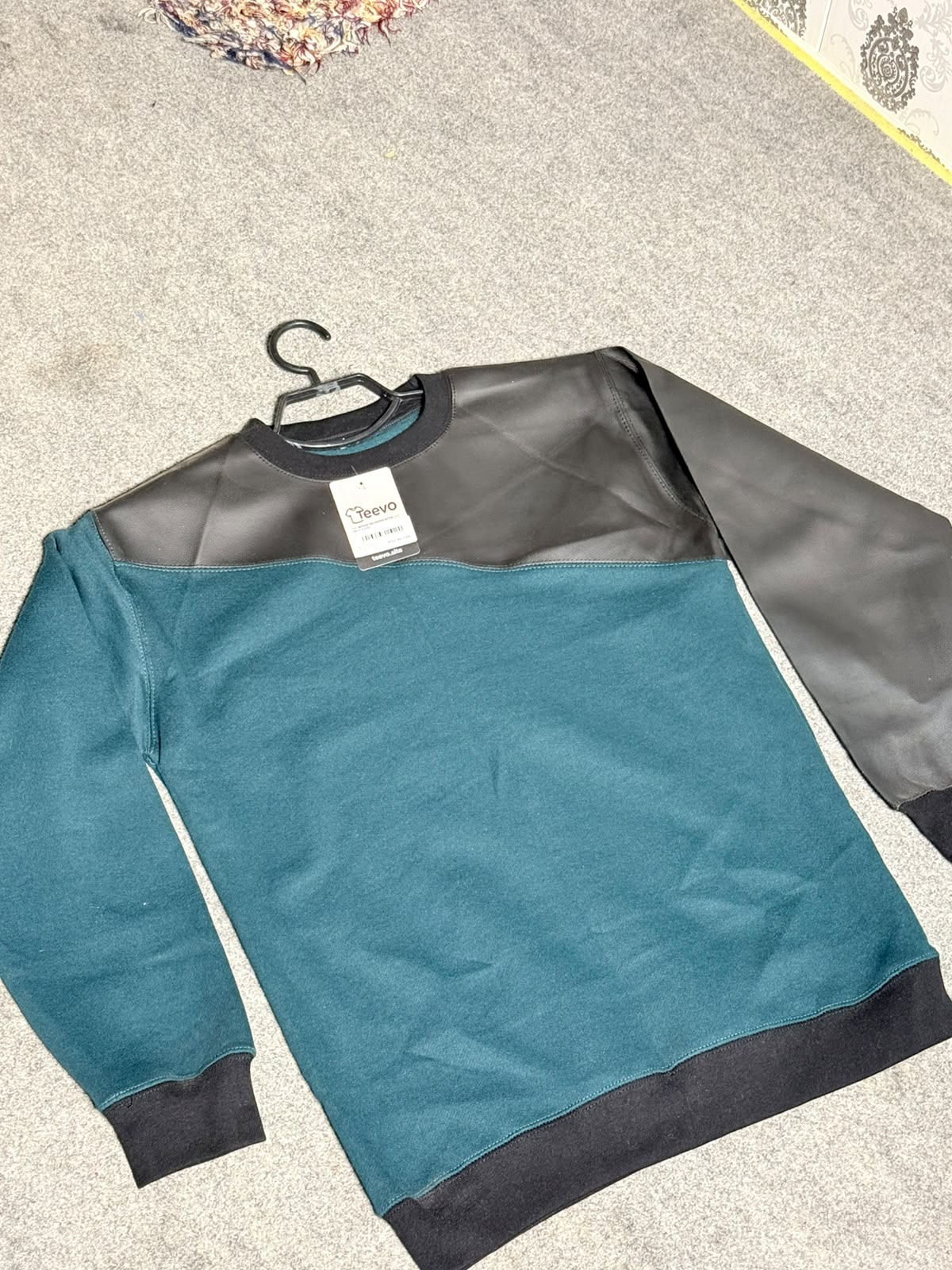 A color-block crewneck sweatshirt with a teal green body, black ribbed collar, cuffs, and hem, and a black faux-leather yoke across the shoulders. The sweatshirt is laid flat on a gray carpeted floor with a black hanger still attached.