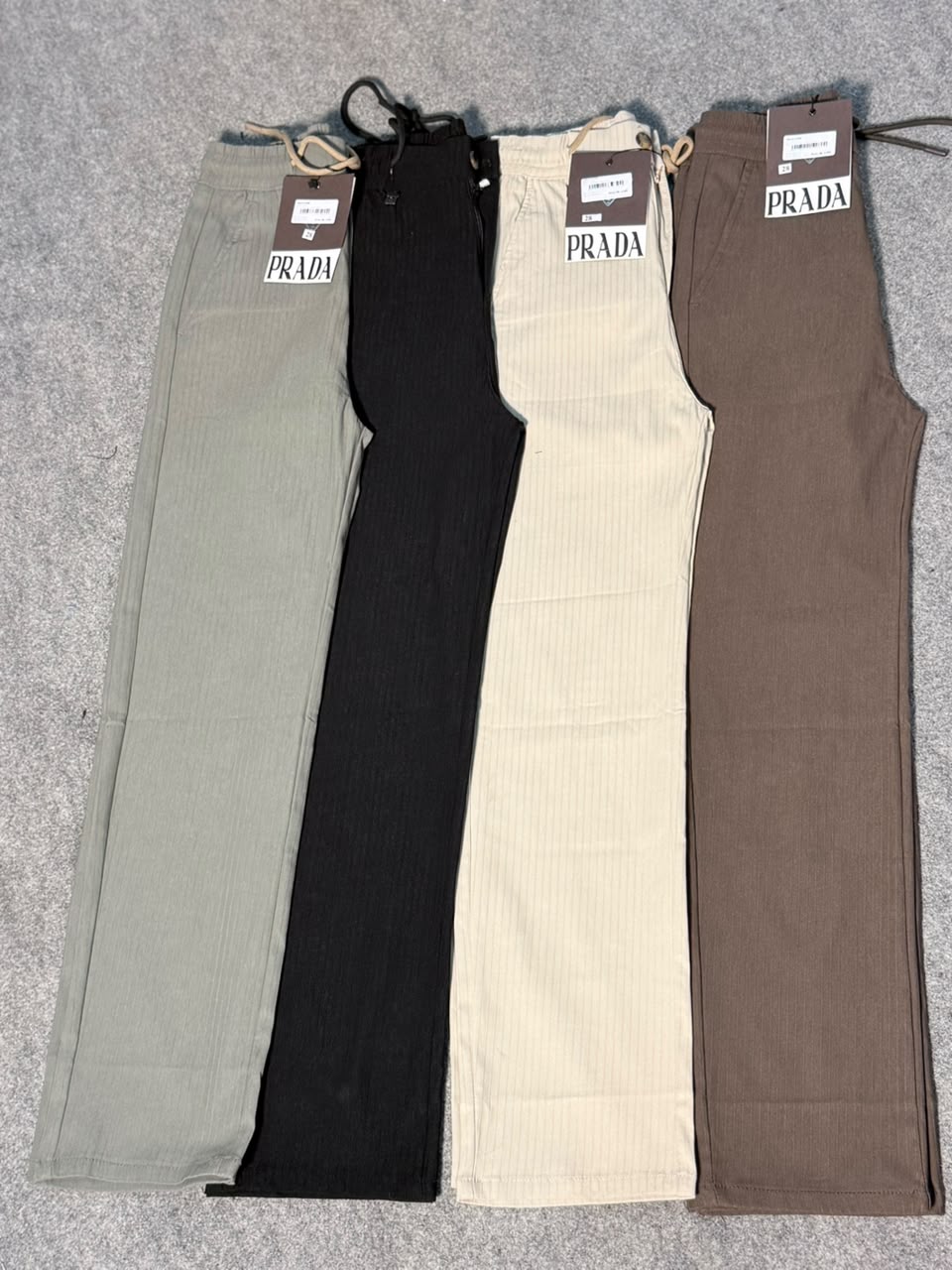 Four Prada ribbed knit pants laid flat on a gray textured surface, shown in sage green, black, cream, and taupe brown.