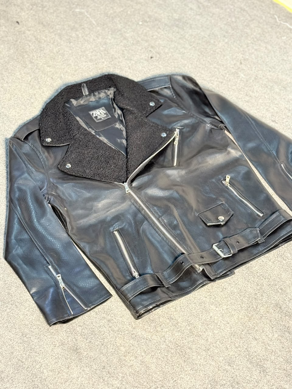 A black faux-leather biker jacket with a black fleece collar, laid flat on a gray concrete surface. The jacket features silver zippers, a front flap pocket, and a matching black belt.