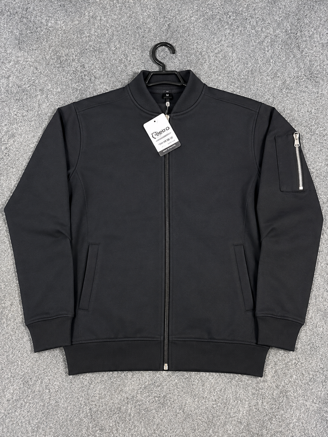A black zip-up bomber jacket with a stand collar, ribbed cuffs and hem, and a zippered sleeve pocket, laid flat on a gray textured background with a hangtag attached.