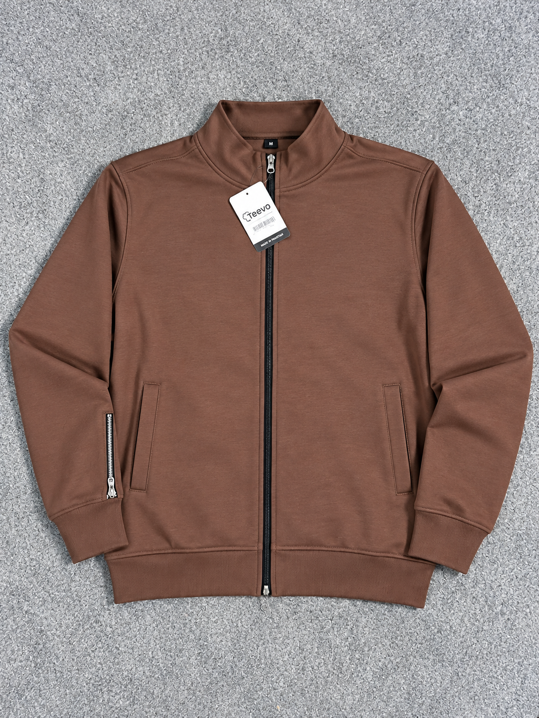A brown zip-up jacket with a stand-up collar, laid flat on a textured gray background. It features a black zipper, side zip pocket, and a hangtag.