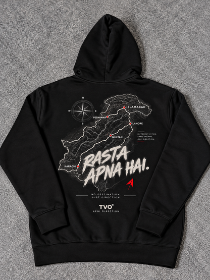 A black pullover hoodie laid flat on a gray textured surface, showing the back side with a large graphic print. The print features a white map outline, a compass rose, and the text 'Rasta Apna Hai.