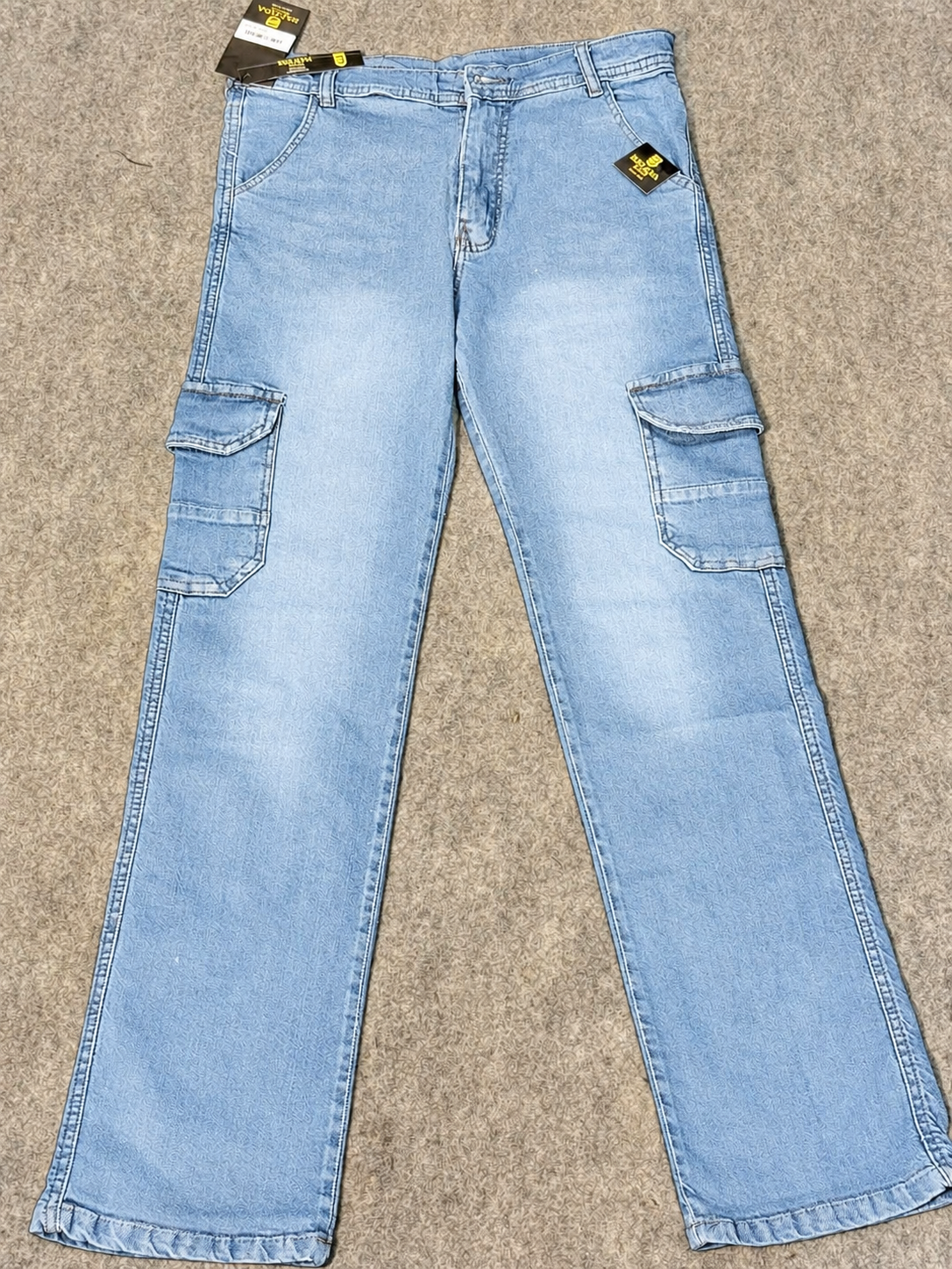 Light blue denim cargo jeans laid flat on a textured gray carpet, showing side cargo pockets and retail tags.