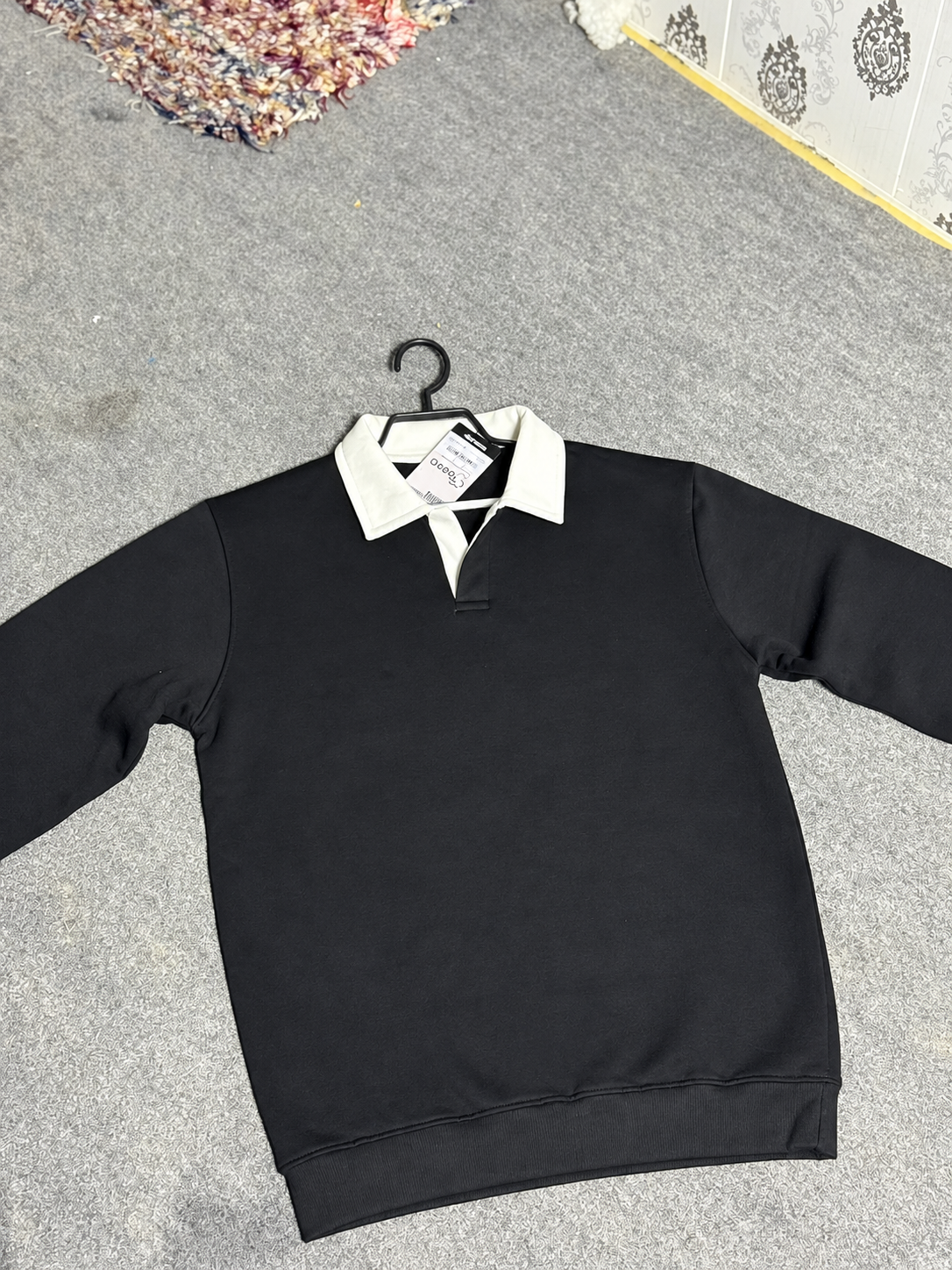 A black long-sleeve sweatshirt with a contrasting white polo-style collar, hanging on a black hanger on a gray carpeted floor.