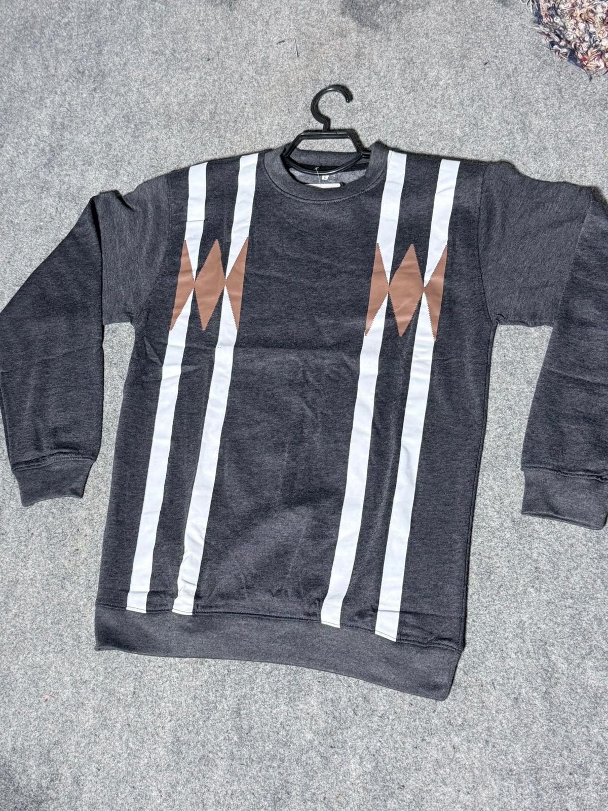A dark gray crewneck sweatshirt with vertical white stripes and brown geometric diamond patterns on the chest, laid flat on a gray textured surface with a black hanger.