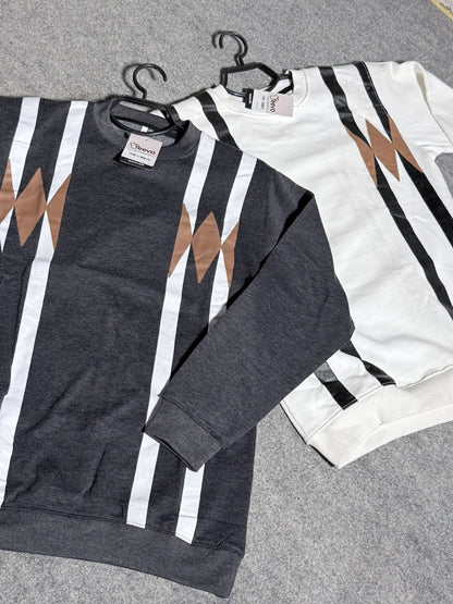 Two crewneck sweatshirts with a geometric stripe pattern, one in dark gray and one in off-white, laid flat on a gray felt surface. Both feature vertical white and black stripes with brown angular shapes on the chest.
