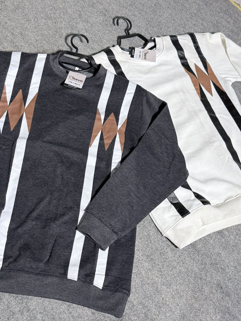 Two crewneck sweatshirts with a geometric stripe pattern, one in dark gray and one in off-white, laid flat on a gray felt surface. Both feature vertical white and black stripes with brown angular shapes on the chest.