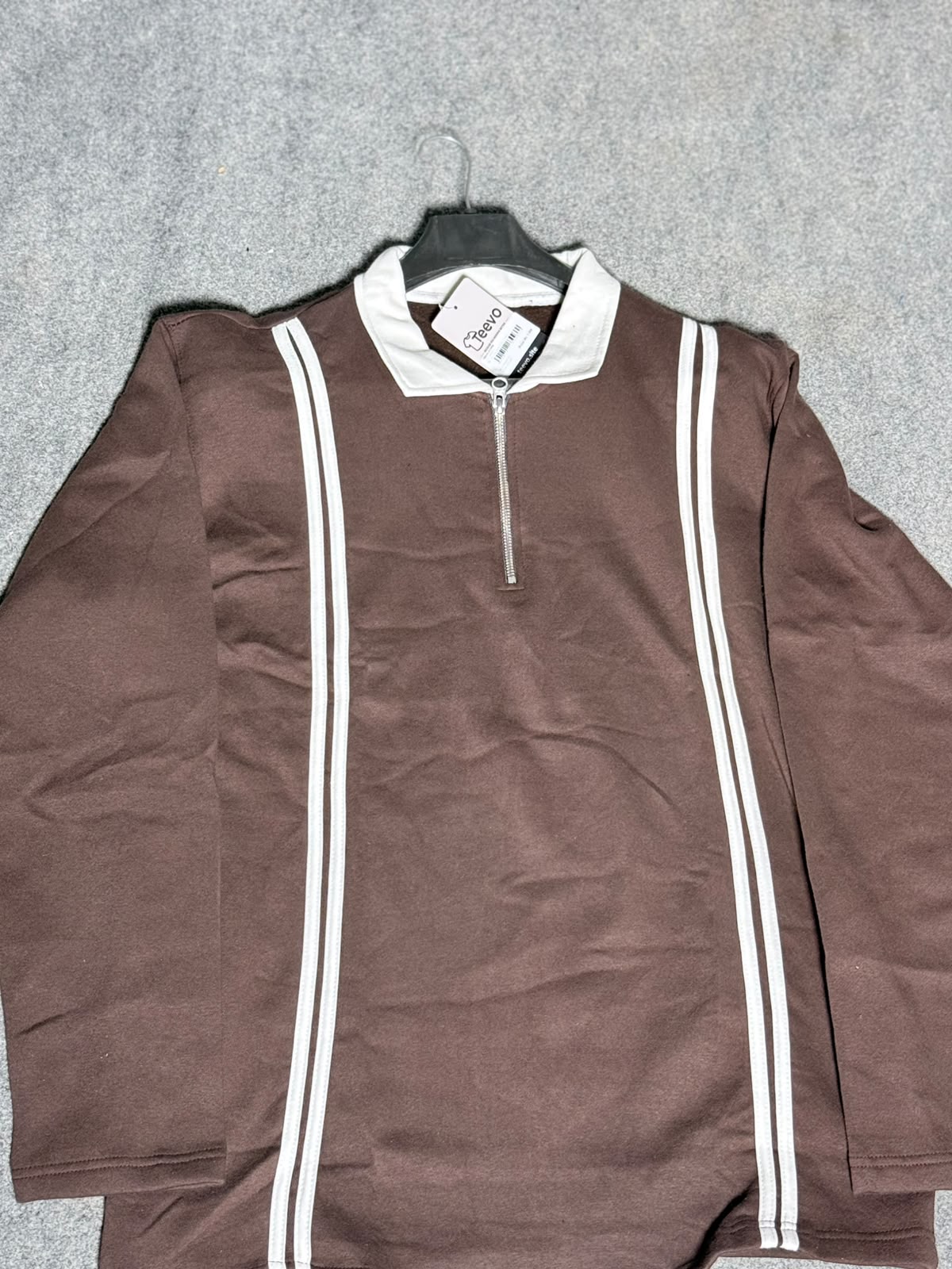 A brown long-sleeve quarter-zip pullover sweatshirt with white double stripes down the front, hanging on a black hanger against a gray textured background.