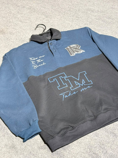 A long-sleeve quarter-zip sweatshirt laid flat on a gray carpet. The top half is slate blue with a dark collar, and the bottom half is dark charcoal gray.