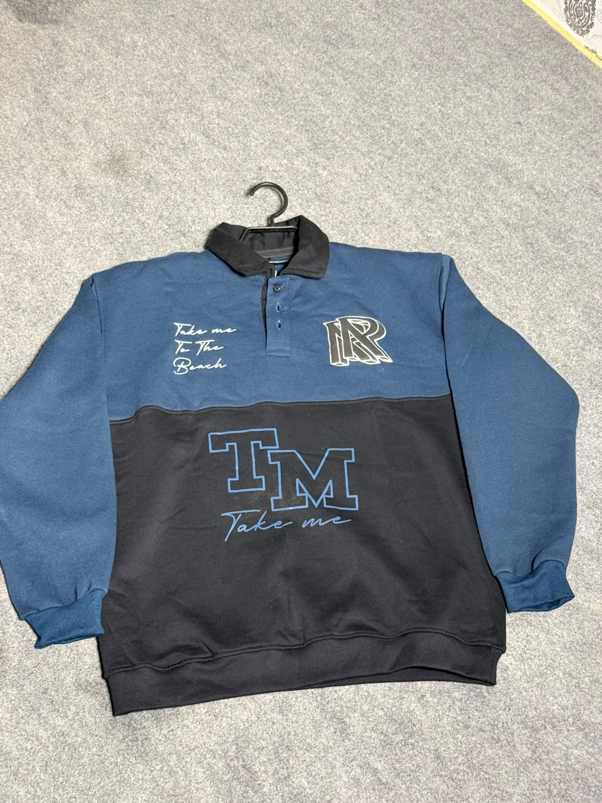 A long-sleeve quarter-zip sweatshirt in blue and black, laid flat on a gray carpeted floor. The top half is blue, the bottom half is black, and it features 'TM Take me' graphics and a stylized 'AR' logo on the chest.