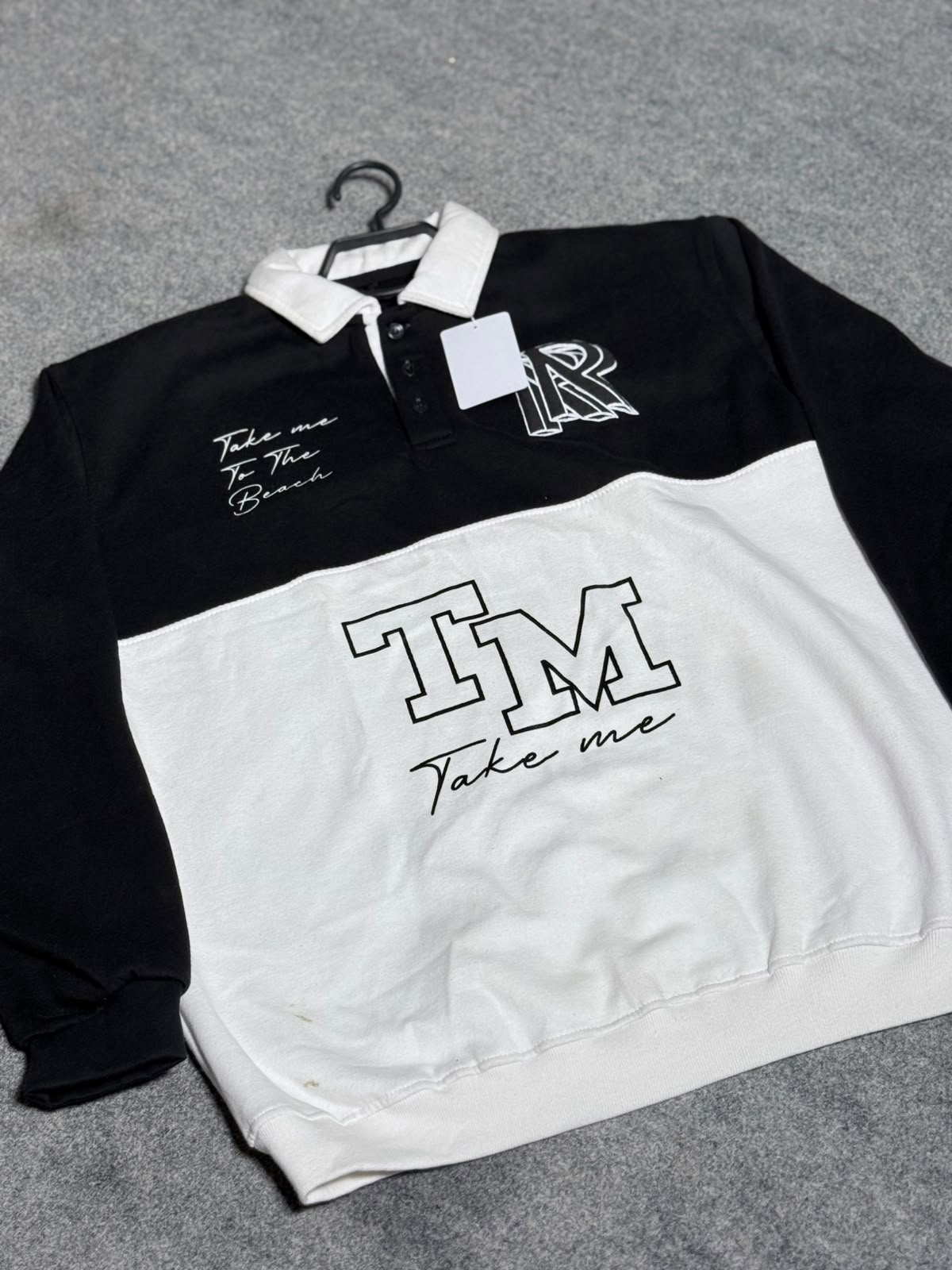A long-sleeve rugby-style shirt in black and white, laid flat on a gray textured surface. The top half is black with a white collar, featuring a white 'R' monogram and 'Take me To The Beach' script on the chest.