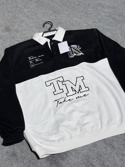A long-sleeve rugby-style shirt in black and white, laid flat on a gray textured surface. The top half is black with a white collar, featuring a white 'R' monogram and 'Take me To The Beach' script on the chest.