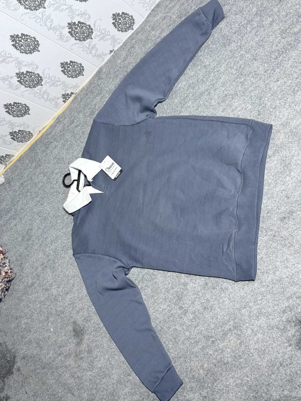A slate blue long-sleeve sweatshirt laid flat on a gray carpeted floor, with white retail tags still attached near the left chest.