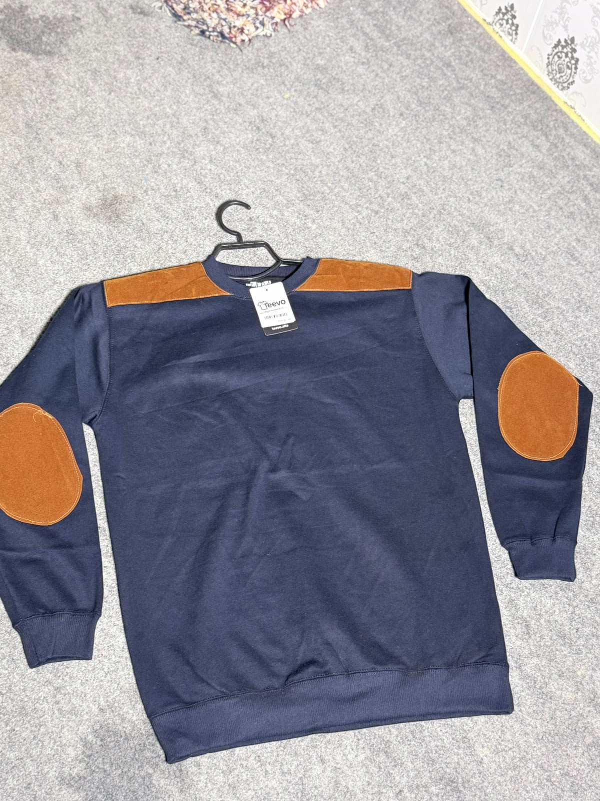 A navy blue crewneck sweatshirt with tan contrast panels across the chest and matching tan oval elbow patches, laid flat on a gray carpeted floor with a black hanger.