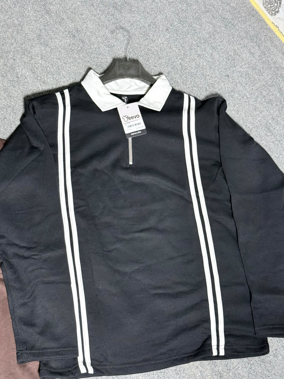 A black long-sleeve rugby-style shirt with white double stripes down the front and a white collar, hanging on a black hanger against a gray textured background.