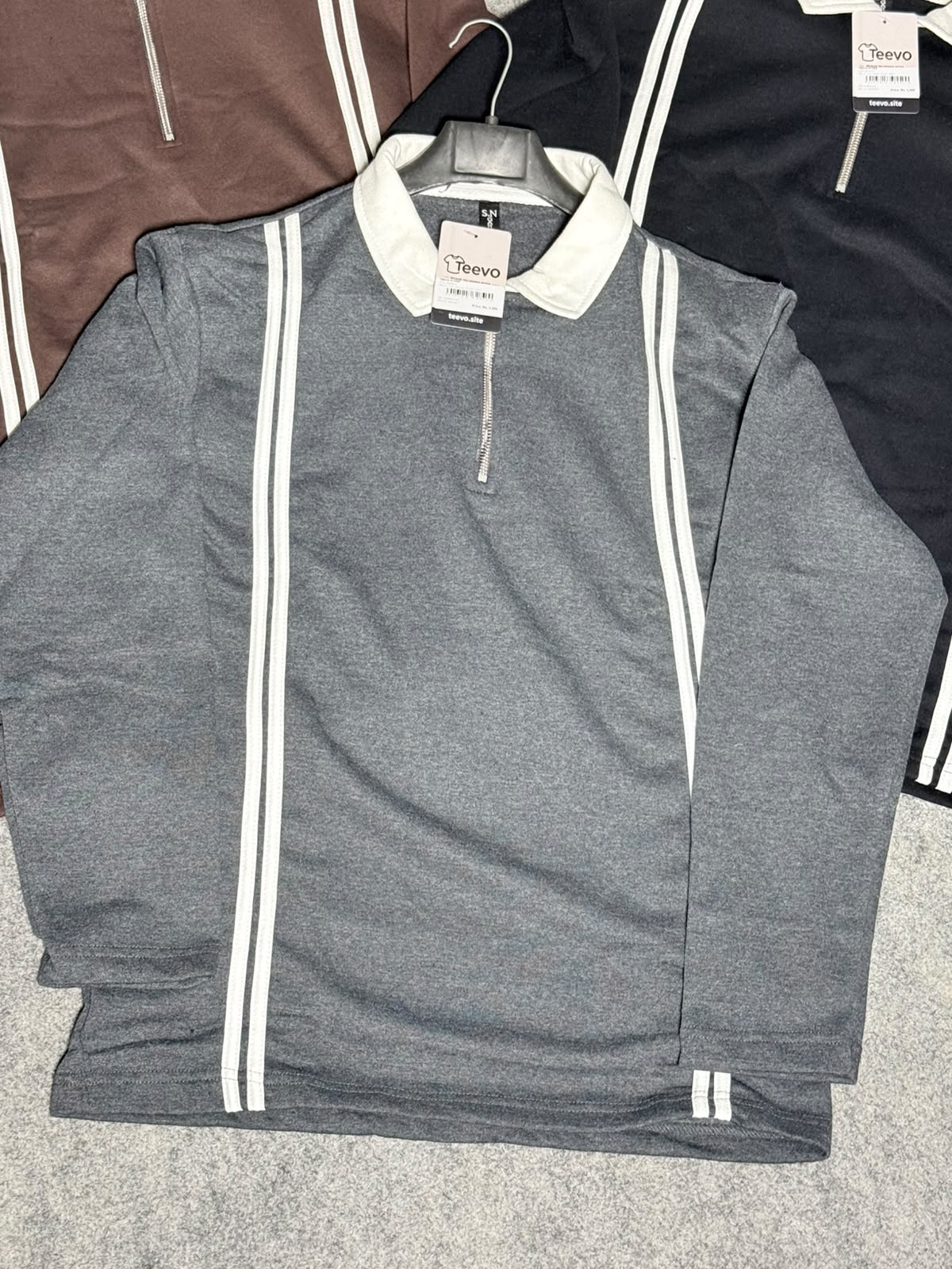 A heather gray long-sleeve quarter-zip pullover with white double stripes down the front and collar, shown on a hanger with other color options partially visible behind it.