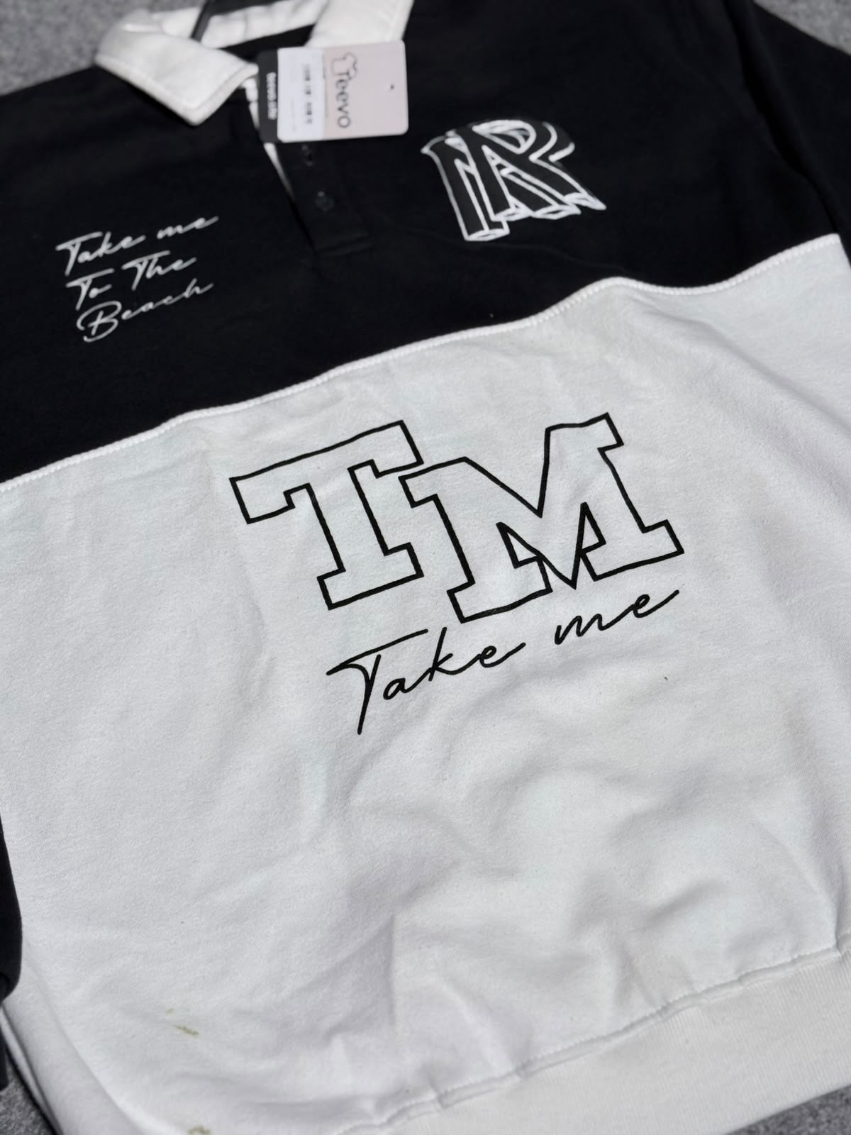 A color-blocked short-sleeve polo shirt with a black top half and white bottom half. The top half features a white collar, a white 'IR' monogram on the left chest, and white script text reading 'Take me To The Beach' on the right chest.