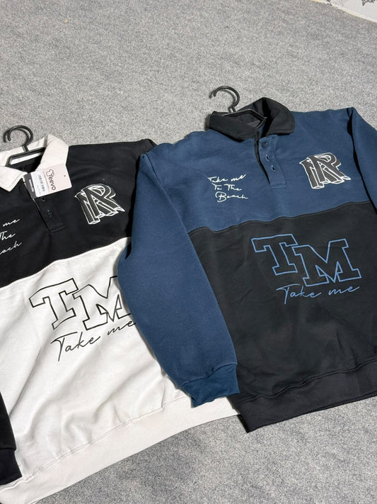 Three color-blocked long-sleeve rugby pullovers with embroidered 'IM' and 'Take me' graphics, displayed on hangers over a gray textured surface. The visible colorways are black and white, blue and black, and navy and black.