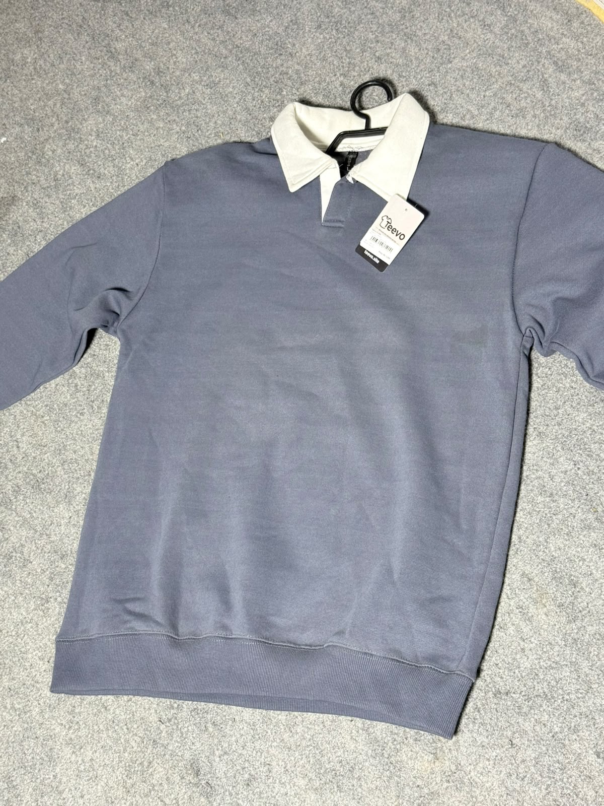 A gray long-sleeve polo shirt with a contrasting white collar, laid flat on a gray carpeted floor with a hangtag still attached.