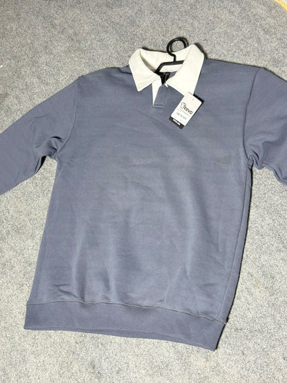 A gray long-sleeve polo shirt with a contrasting white collar, laid flat on a gray carpeted floor with a hangtag still attached.