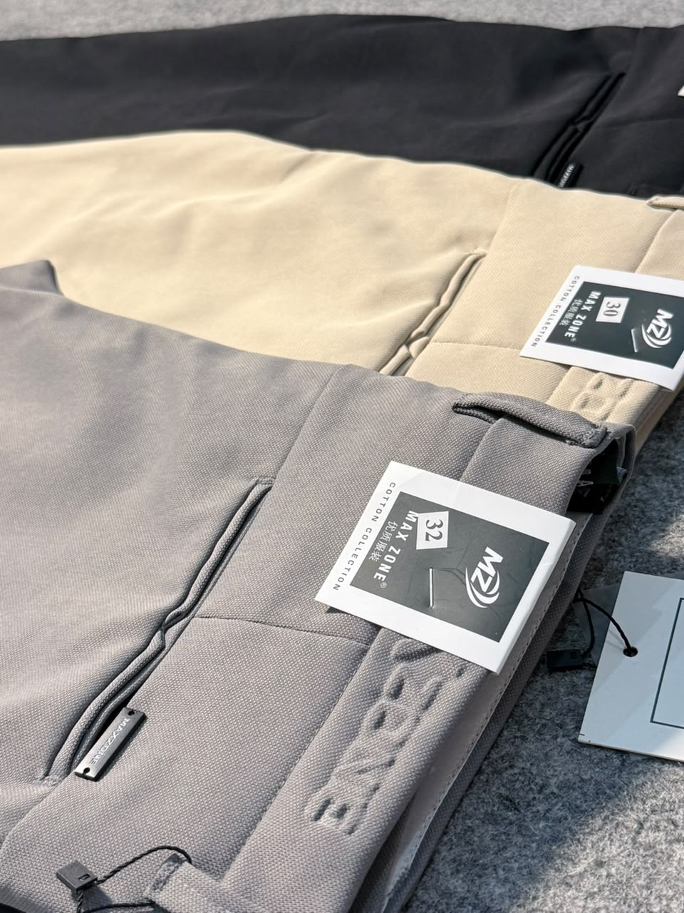 A close-up of folded gray pants with a visible hangtag, placed over beige and black pants on a textured gray surface. The gray pants show an embossed logo and a zip pocket.