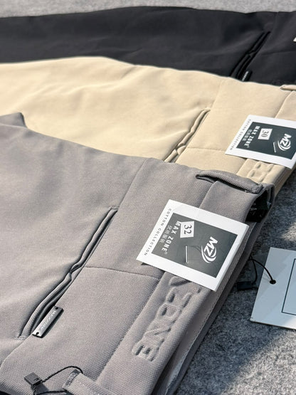 A close-up of folded gray pants with a visible hangtag, placed over beige and black pants on a textured gray surface. The gray pants show an embossed logo and a zip pocket.