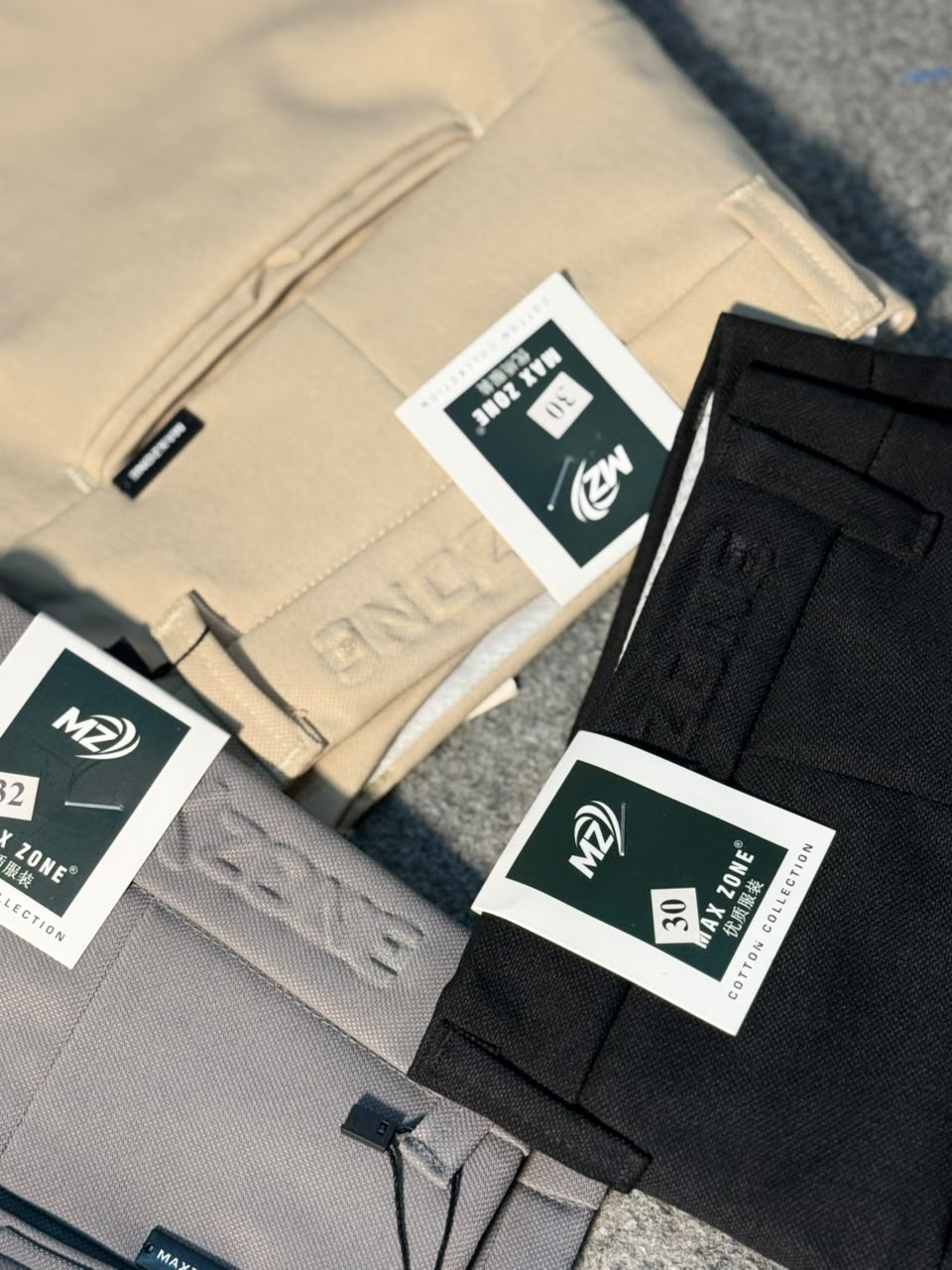 Three pairs of folded pants from the M21 'AT ZONE' collection are arranged diagonally on a gray textured surface. The pants are in beige, black, and gray, each featuring a white brand tag with black text and a small black logo.
