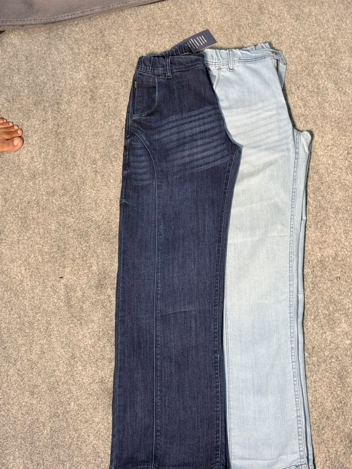Two pairs of folded jeans laid flat on a gray carpet: one pair in dark blue denim and one pair in light blue denim.