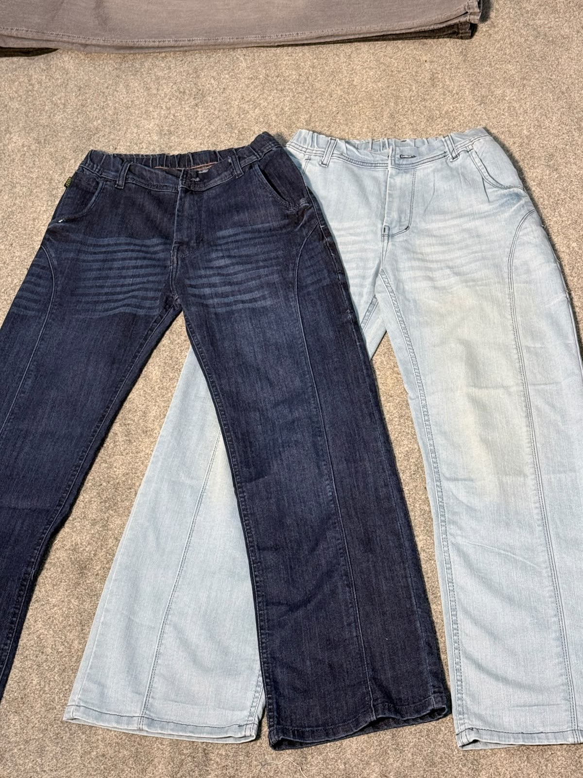 Two pairs of jeans laid flat on a beige carpet: one dark blue denim and one light blue denim, both shown from the front.