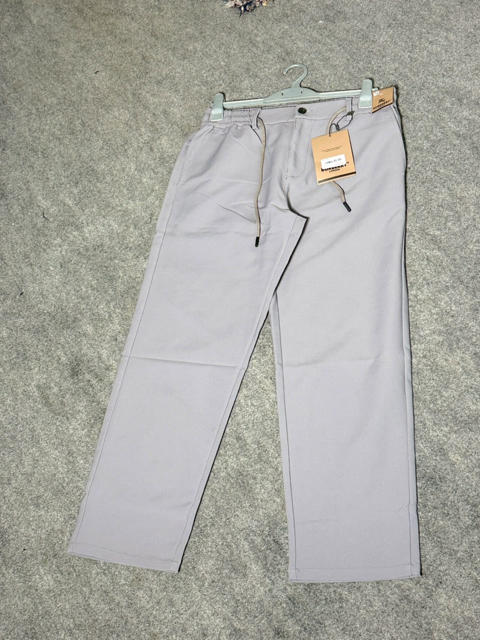 A pair of light gray drawstring trousers laid flat on a gray carpeted floor, hanging from a metal hanger with a paper brand tag attached.