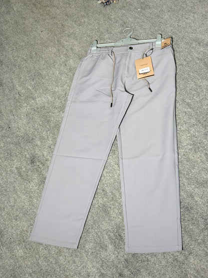A pair of light gray drawstring trousers laid flat on a gray carpeted floor, hanging from a metal hanger with a paper brand tag attached.