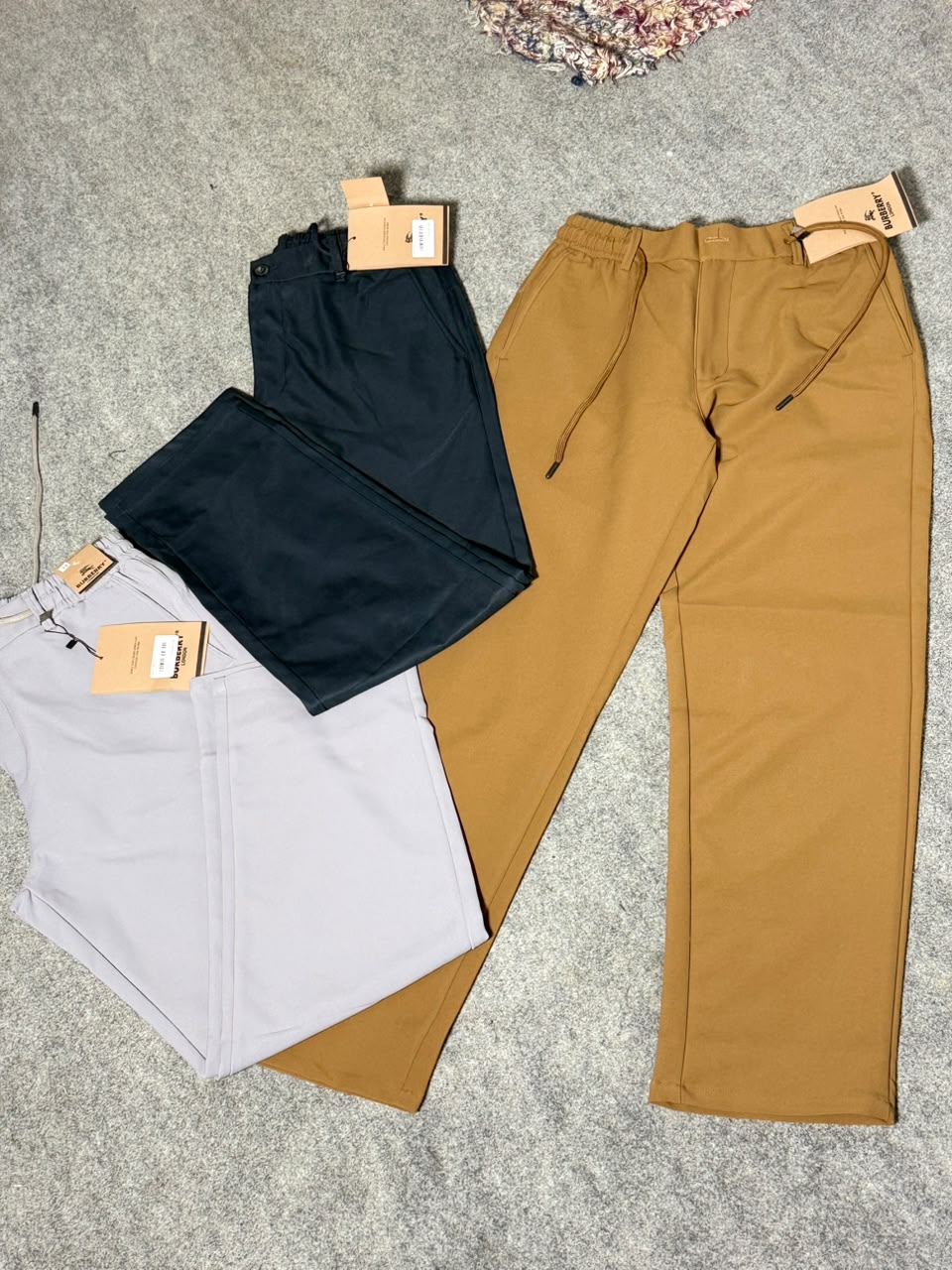 Three pairs of folded drawstring pants laid out on a gray textured surface: one tan, one dark navy, and one light gray, each with a paper tag attached.