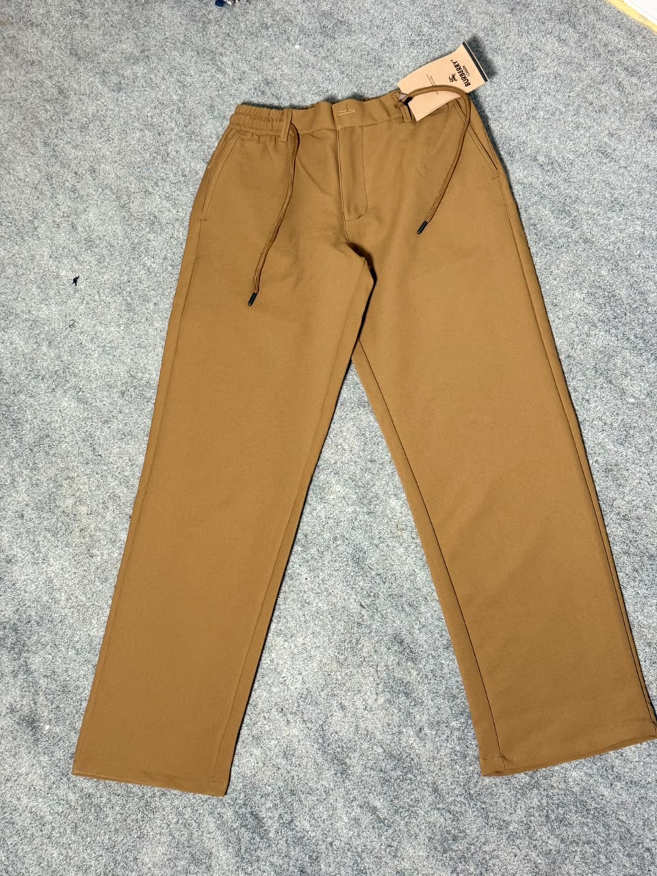 A pair of solid brown pants with a drawstring waist and a paper tag attached near the hem, laid flat on a gray textured carpet.