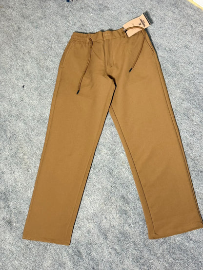 A pair of solid brown pants with a drawstring waist and a paper tag attached near the hem, laid flat on a gray textured carpet.