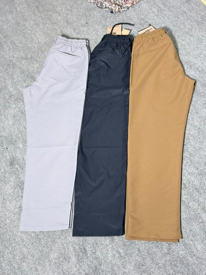 Three pairs of casual drawstring pants laid flat on a gray textured surface, shown in light gray, dark navy, and tan.