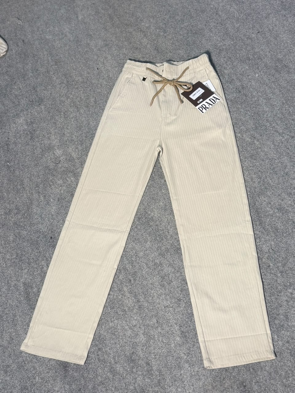 Prada cream-colored corduroy pants with a drawstring waist and a hanging brand tag, laid flat on a gray textured surface.