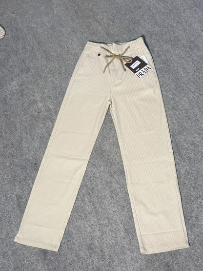 Prada cream-colored corduroy pants with a drawstring waist and a hanging brand tag, laid flat on a gray textured surface.