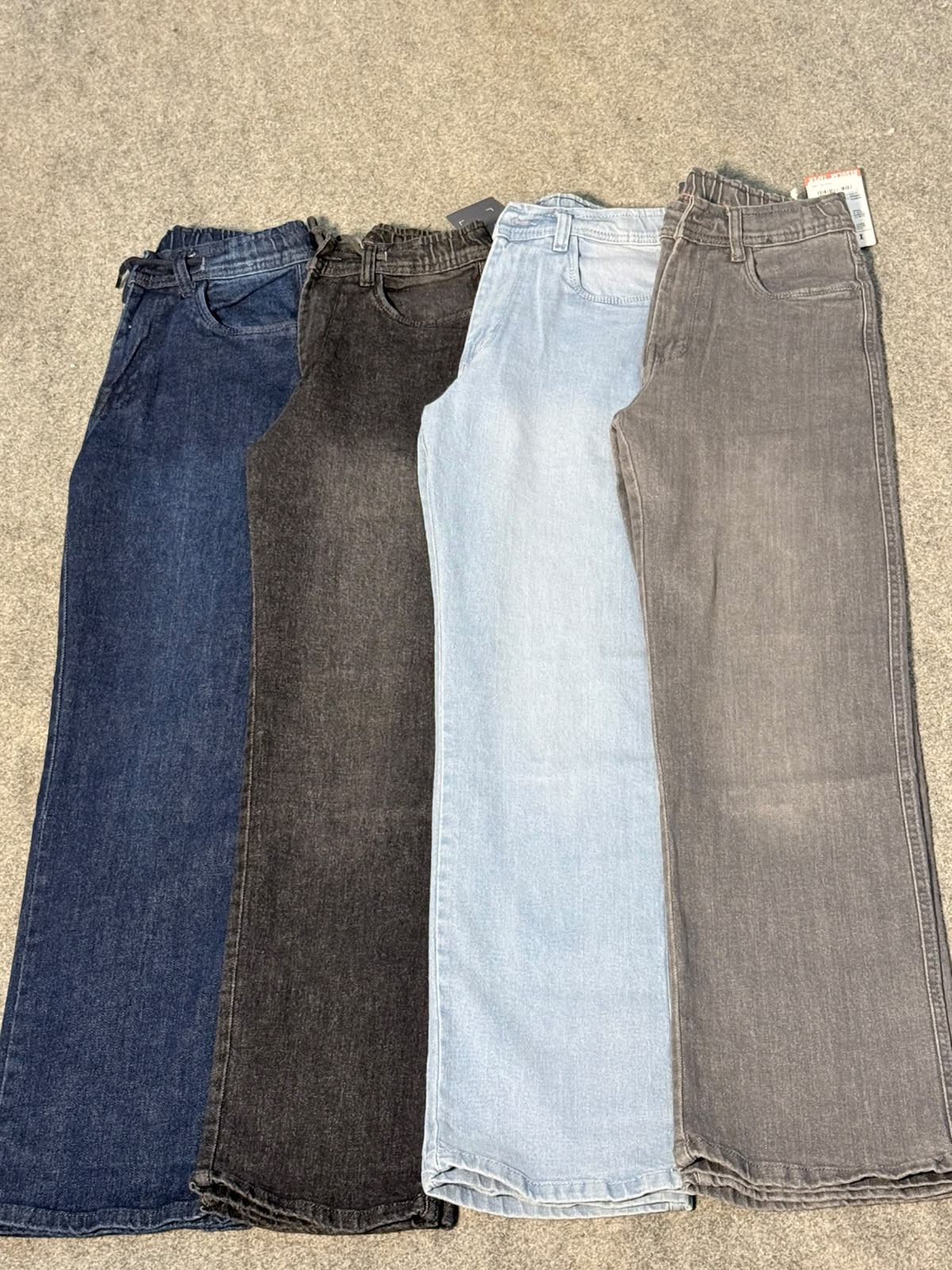 Four pairs of wide-leg jeans in different denim washes laid flat on a gray carpet: dark blue, faded black, light blue, and medium gray.