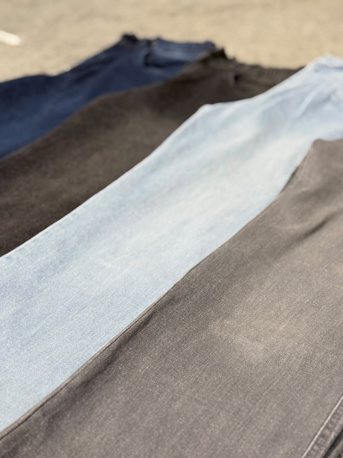 Four pairs of folded jeans laid out diagonally in a row, showing different denim washes: dark blue, charcoal gray, light blue, and medium gray, with a concrete ground background.