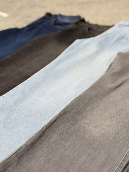 Four pairs of folded jeans laid out diagonally in a row, showing different denim washes: dark blue, charcoal gray, light blue, and medium gray, with a concrete ground background.