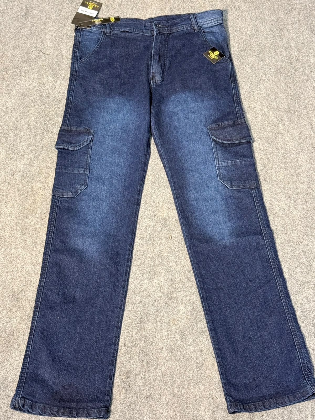 A pair of medium-wash blue denim cargo jeans laid flat on a beige carpet background, featuring side cargo pockets and a small yellow embroidered logo patch on the upper right hip.