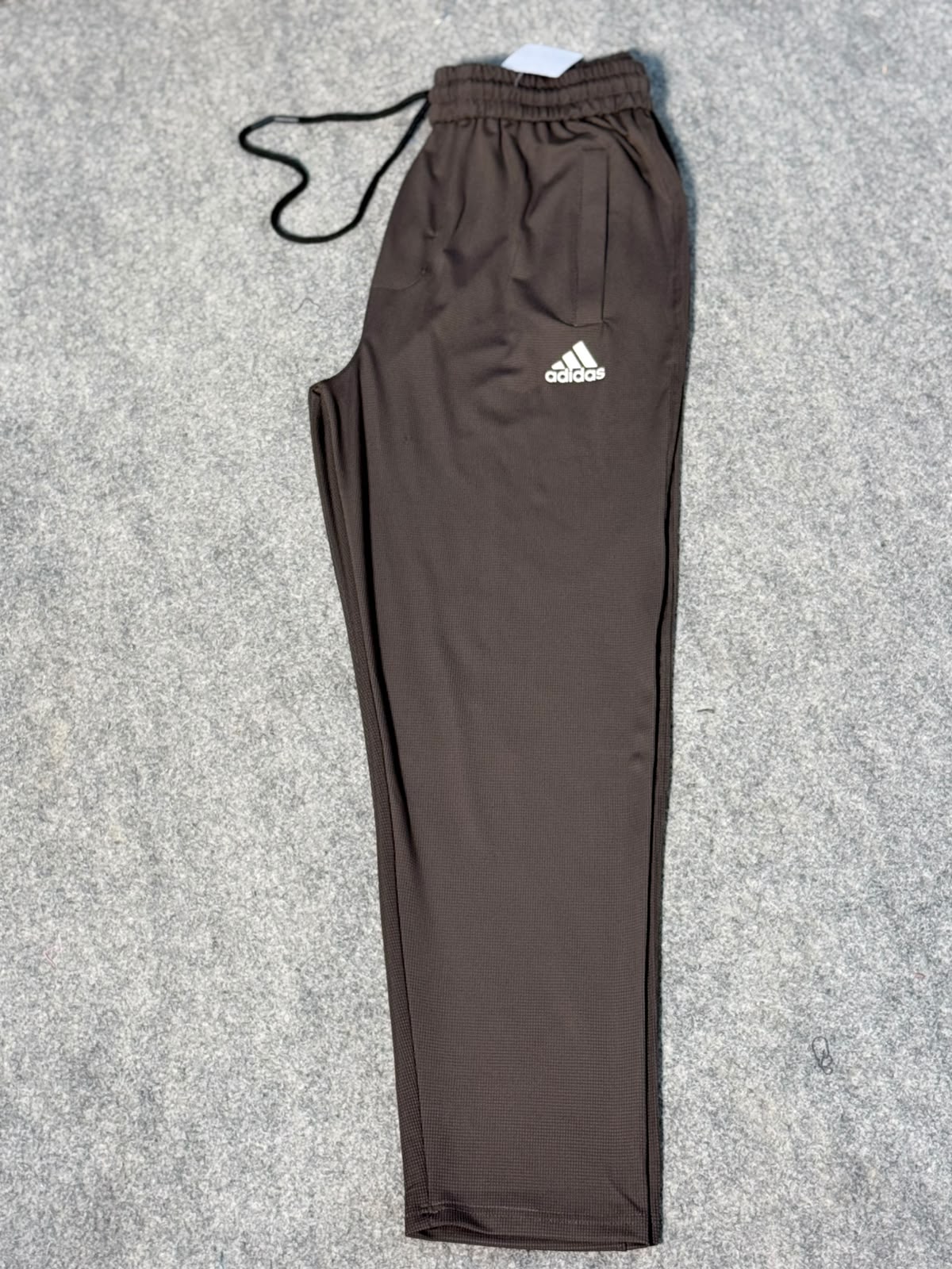 A pair of dark brown Adidas track pants with a black drawstring and a white logo on the left thigh, laid flat on a gray textured background.