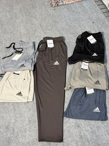 Six pairs of Adidas athletic shorts in various colors (gray, black, beige, brown, light gray, and navy) are neatly folded and arranged on a gray carpeted floor. Each pair has its white Adidas logo and brand tag visible.