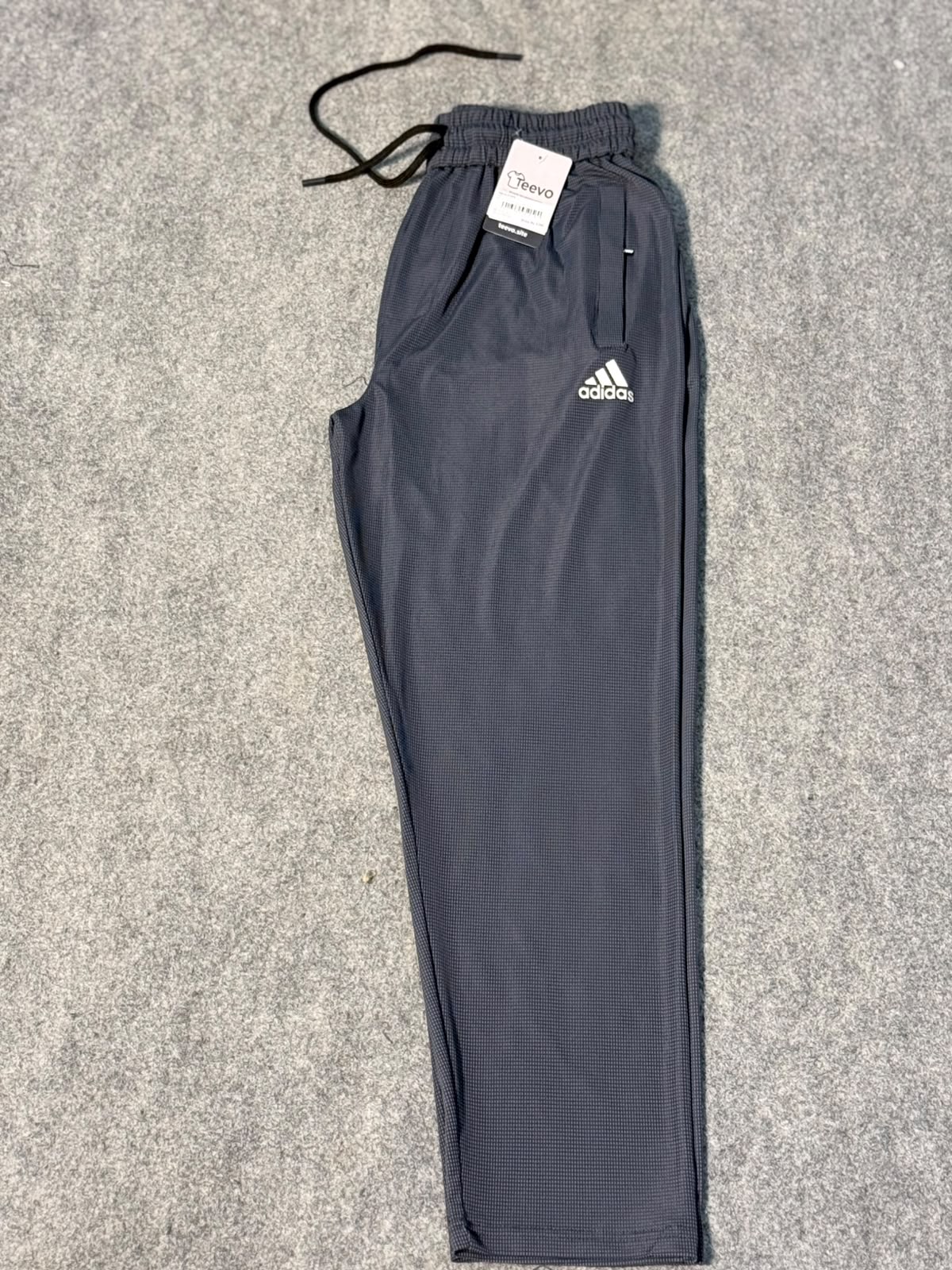 A pair of dark gray Adidas athletic pants with a white logo on the left thigh, laid flat on a gray textured carpet. The pants have an elastic waistband with a black drawstring and a white retail tag attached.