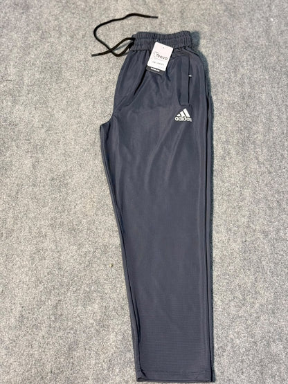A pair of dark gray Adidas athletic pants with a white logo on the left thigh, laid flat on a gray textured carpet. The pants have an elastic waistband with a black drawstring and a white retail tag attached.