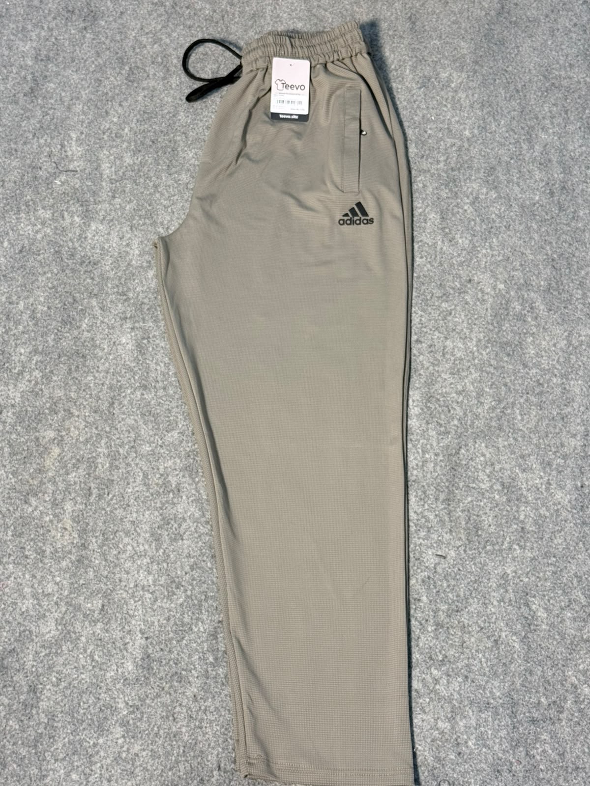 A pair of Adidas jogger sweatpants in a light khaki-beige color, laid flat on a gray textured background. The pants feature an elastic waistband with a black drawstring, a zippered pocket on the left thigh, and the black Adidas logo on the right thigh.