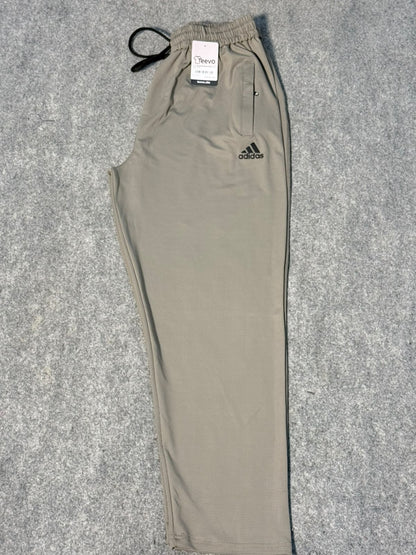 A pair of Adidas jogger sweatpants in a light khaki-beige color, laid flat on a gray textured background. The pants feature an elastic waistband with a black drawstring, a zippered pocket on the left thigh, and the black Adidas logo on the right thigh.