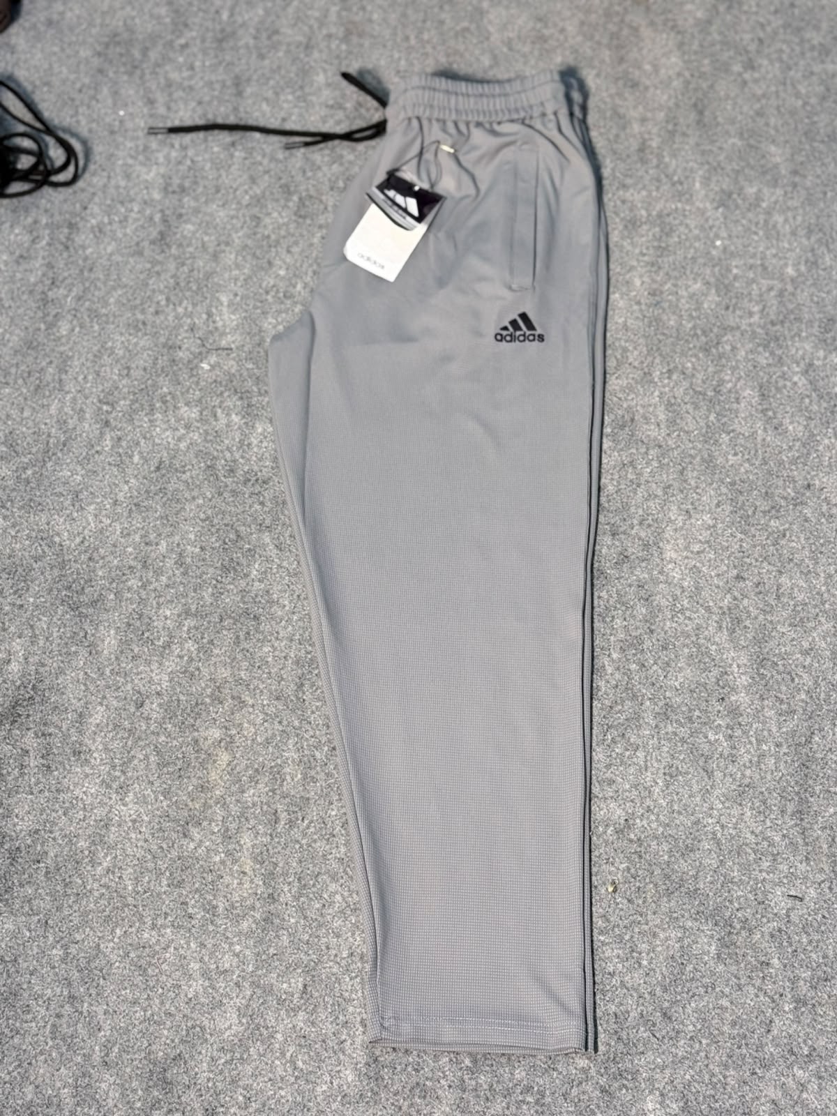 A pair of light gray Adidas jogger pants laid flat on a gray textured surface, with the black drawstring and white hangtag still attached. The black Adidas logo is visible on the upper left thigh.