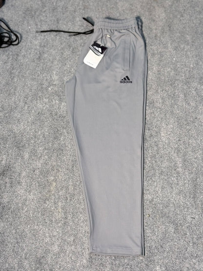 A pair of light gray Adidas jogger pants laid flat on a gray textured surface, with the black drawstring and white hangtag still attached. The black Adidas logo is visible on the upper left thigh.