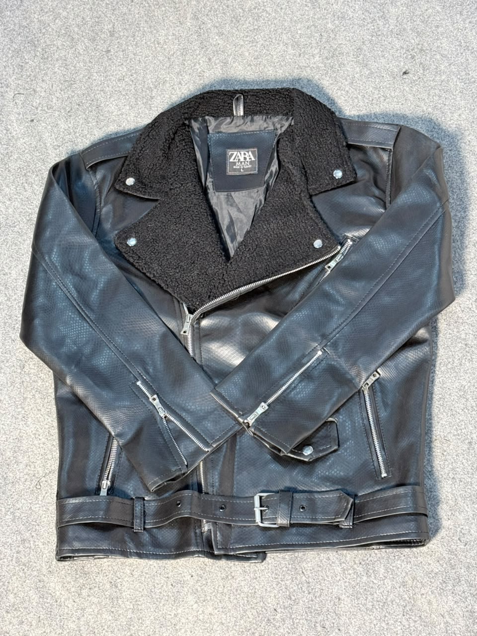 A black leather motorcycle jacket with a black fleece collar, laid flat on a gray textured surface. It features an asymmetrical zipper, zippered chest pockets, and a matching leather belt with a silver buckle.